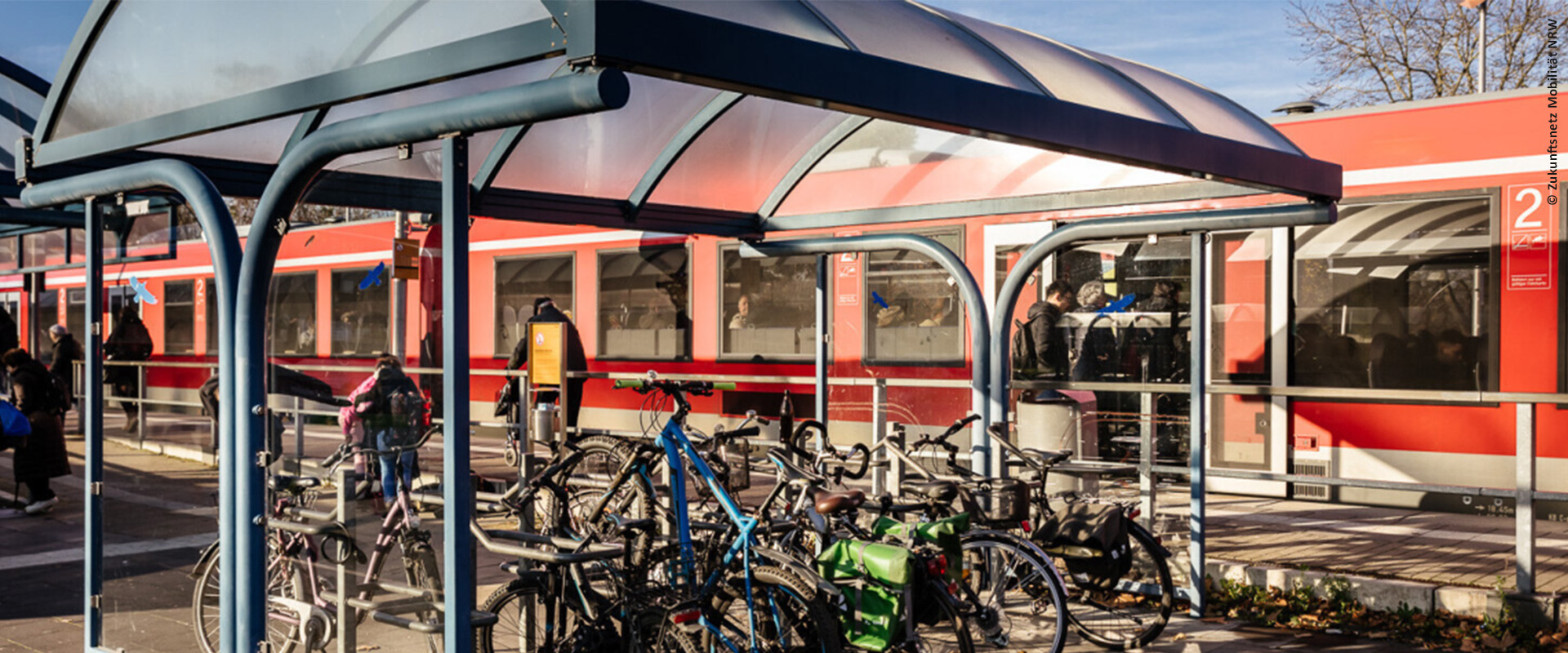 An einem Bahnsteig steht ein roter Zug, Fahrgäste steigen ein, im Vordergrund stehen Fahrräder unter einem Dach