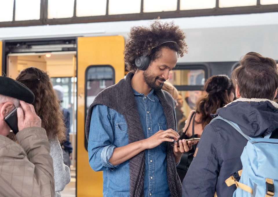 Personen vor einem Zug, in der Mitte ein Mann mit Kopfhörer, der auf sein Smartphone blickt