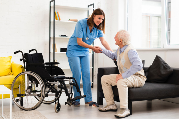 Eine Pflegerin hilft einer Seniorin von der Couch in einen Rollstuhl