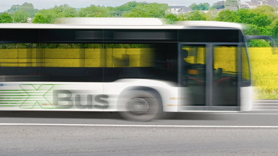 vorderer Teil eines XBusses auf einer Landstraße in der Seitenansicht, im Hintergrund sind Felder und Bäume zu sehen