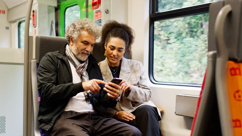 Ein Mann und eine Frau sitzen in einer S-Bahn nebeneinander und schauen auf ein Smartphone