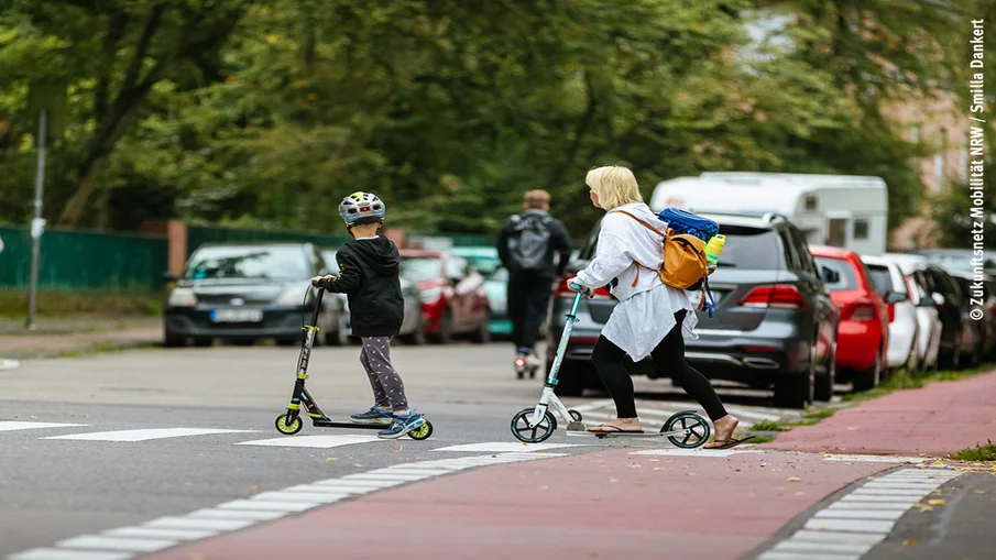 Ein Kind und eine erwachsene Person rollen mit einem Tretroller über einen Zebrastreifen