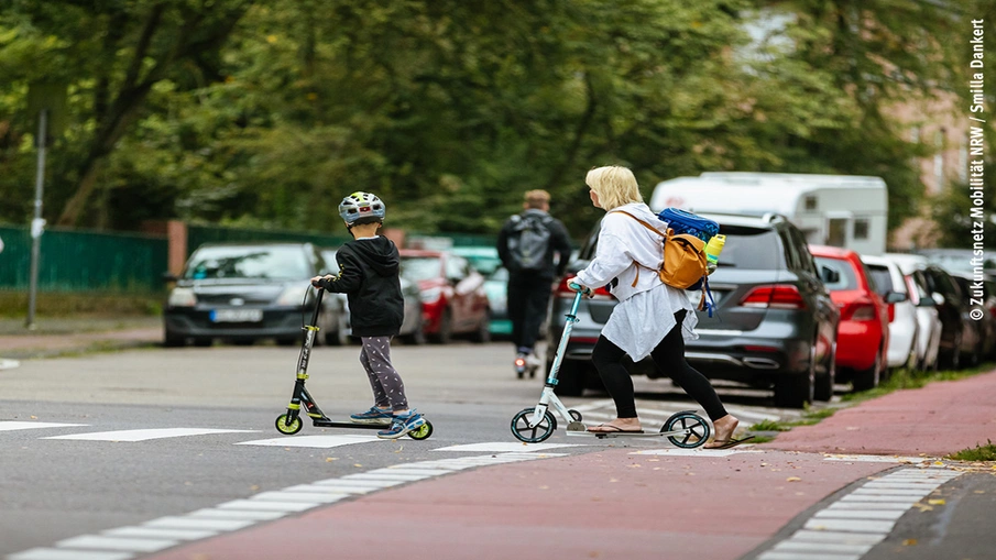 Ein Kind und eine erwachsene Person rollen mit einem Tretroller über einen Zebrastreifen