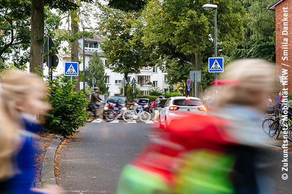 Blick auf eine Straße mit regem Straßenverkehr, Autos warten an einem Zebrastreifen, über den Kinder und Erwachsene laufen oder ihr Fahrrad schieben. Im Vordergrund sind zwei Schulkinder zu sehen