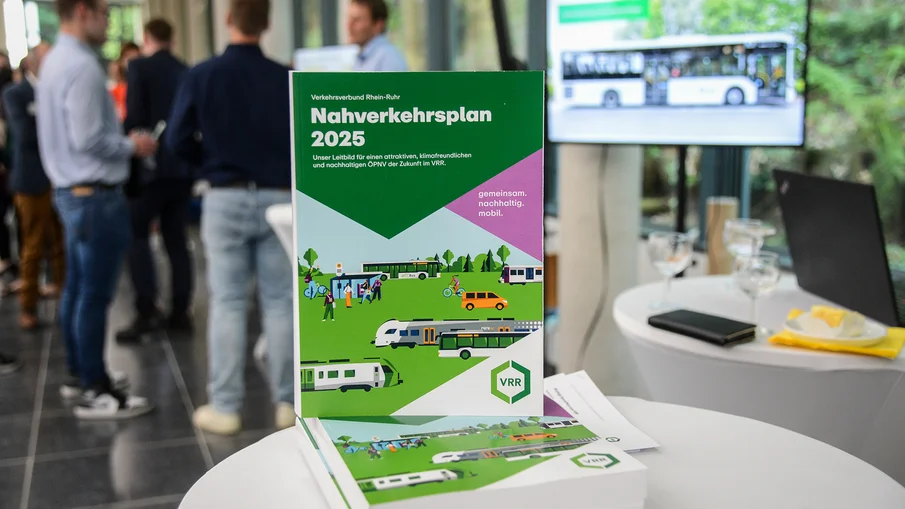 Der VRR-Nahverkehrsplan 2025 auf einem Tisch, im Hintergrund sind Menschen zu sehen