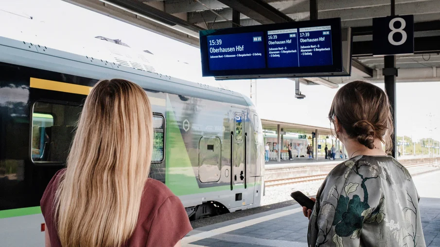 Zwei Personen stehen auf einem Bahnsteig und blicken auf einen Zugzielanzeiger. Am Bahnsteig steht ein Zug des Eisenbahnverkehrsunternehmens Vias