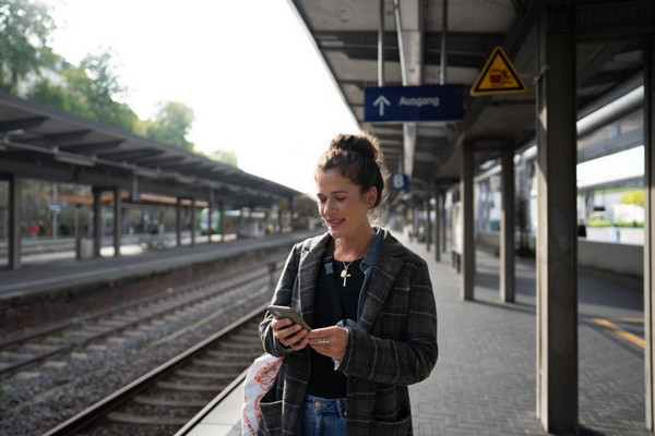 Frau mit Handy in der Hand steht am Bahngleis.