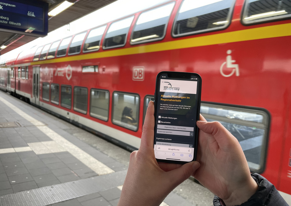 Ein Handy mit der Website zuginfo.nrw, im Hintergrund ein roter Zug