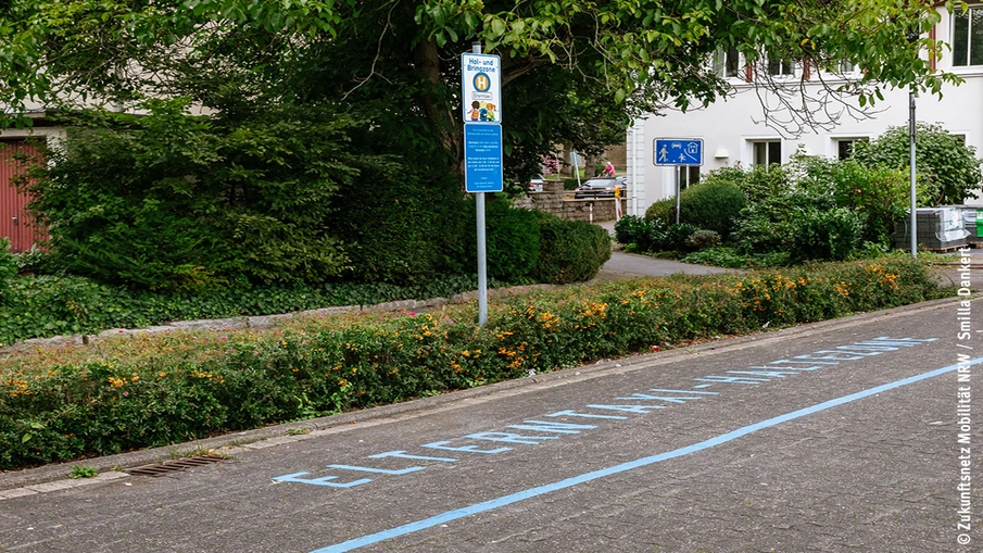 Blick auf eine "Elternhaltestelle", auf dem Boden ist der Schriftzug "Elterntaxi-Haltestelle" zu lesen