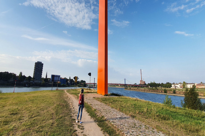 Eine Frau steht an der orange lackierten Stahl-Stele, die den Mündungsbereich der Ruhr in Duisburg markiert
