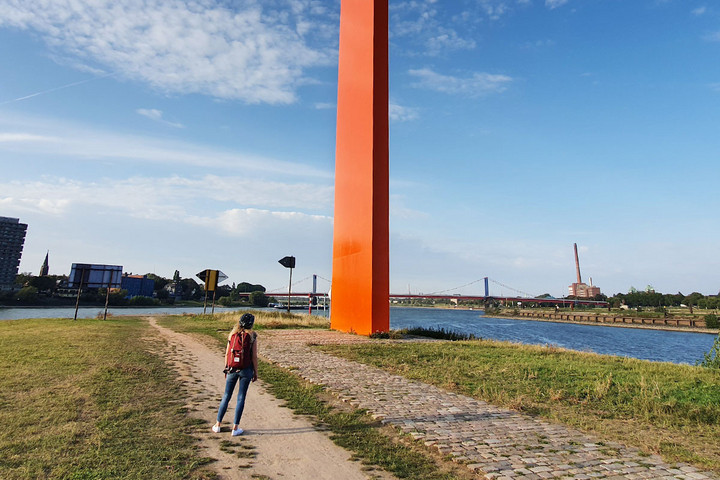 Eine Frau steht an der orange lackierten Stahl-Stele, die den Mündungsbereich der Ruhr in Duisburg markiert
