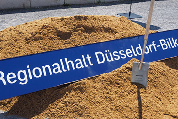 Das Bahnhofsschild von Düsseldorf-Bilk liegt im Sand, daneben ein Spaten
