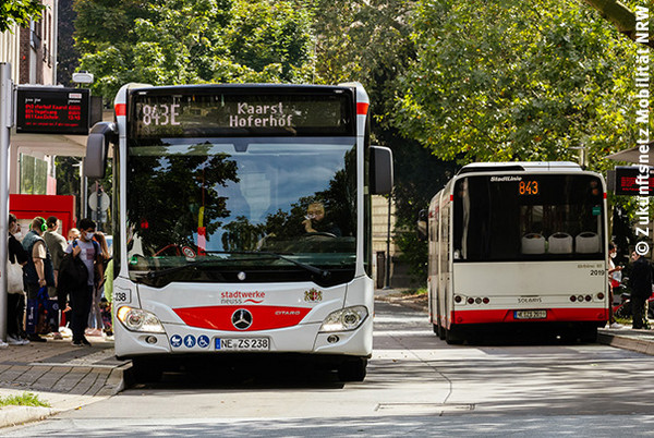Zwei Busse stehen an Haltestellen, Fahrgäste steigen ein und aus