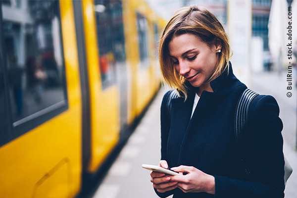 Eine Frau steht an einem Stadtbahn-Bahnsteig und schaut auf ihr Smartphone