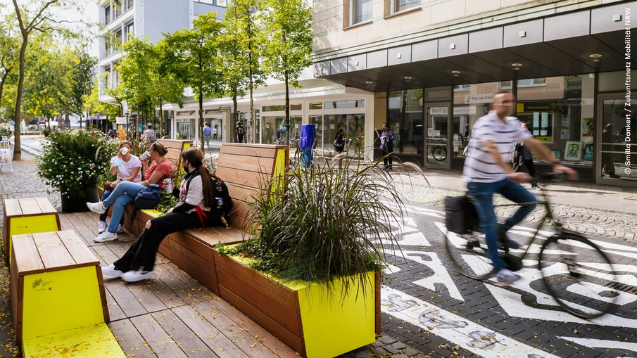 Stadtterrassen in einer Straße, Menschen sitzen auf den Outdoormöbeln, rechts im Bild ist ein Fahrradfahrer zu sehen