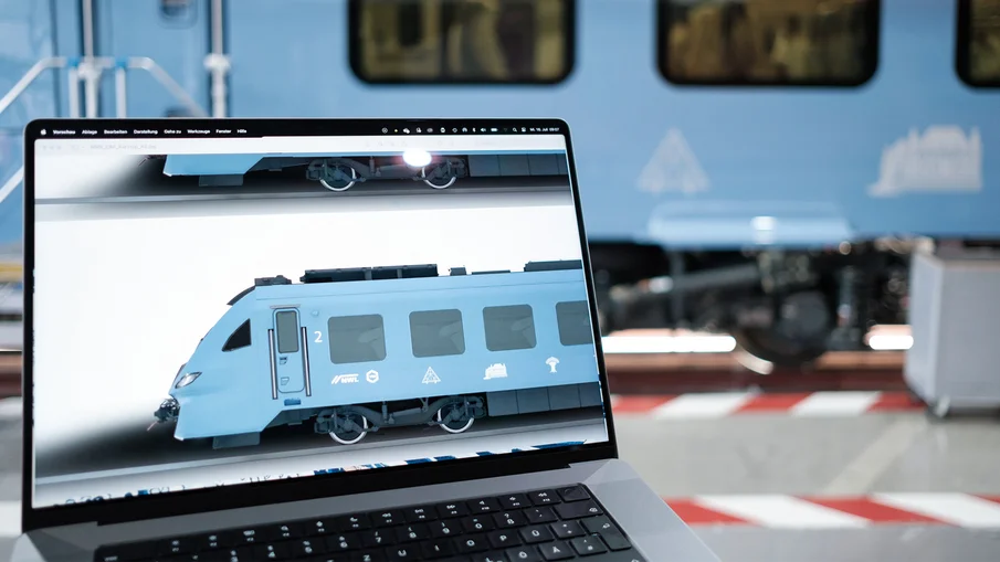 Ein Laptopbildschirm zeigt einen BEMU-Zug, im Hintergrund ist ebenfalls der Zug zusehen.