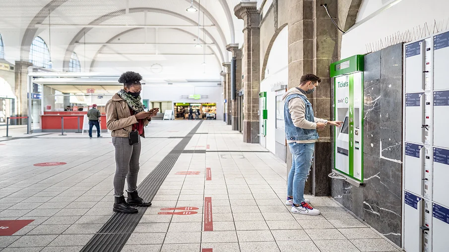 Bahnhofsgebäude von innen, zwei Fahrgäste hintereinander beim Ticketkauf an einem Automaten