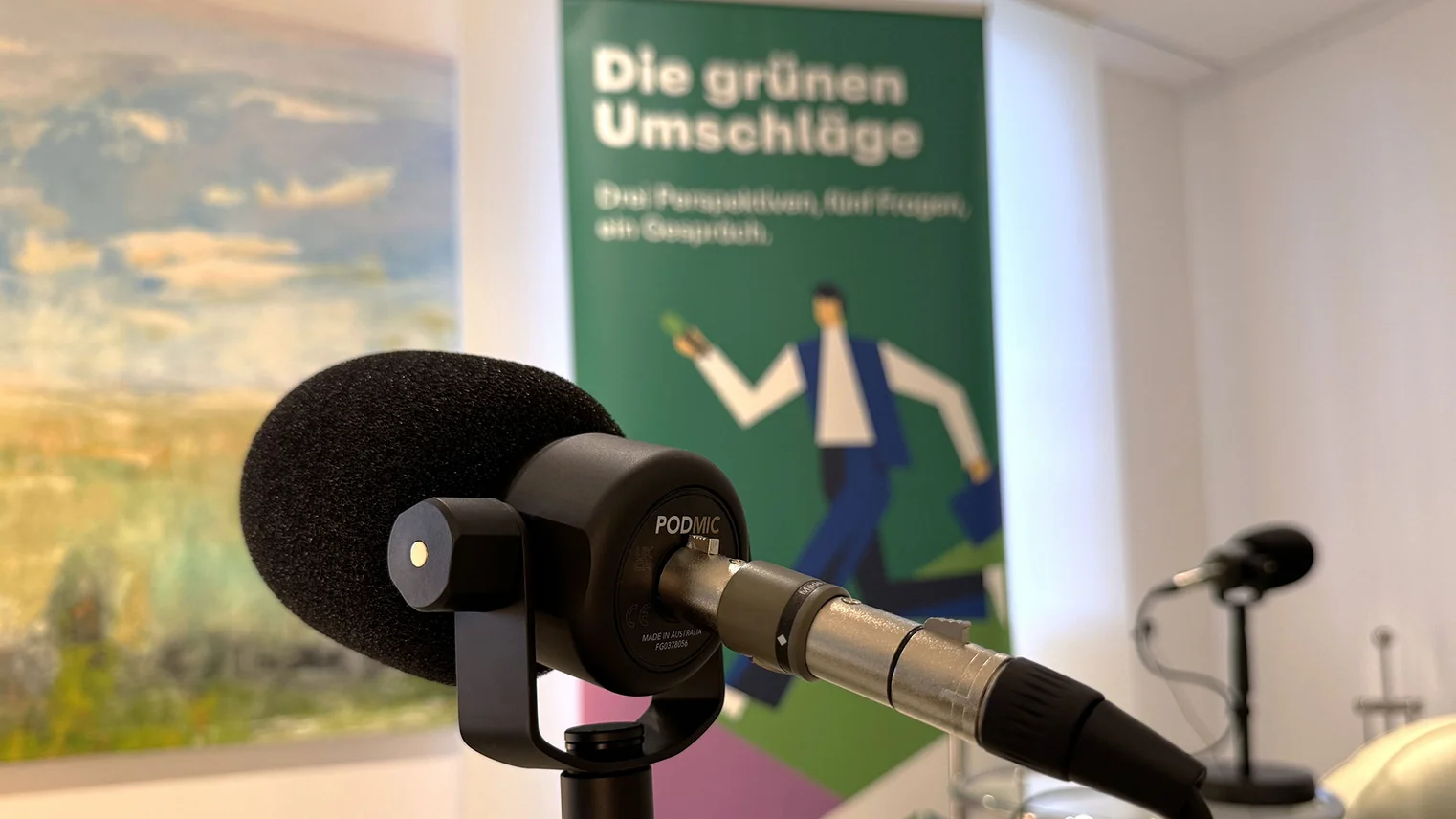 © VRR VRR-Podcast "Die grünen Umschläge": Blick auf ein Mikrofon, im Hintergrund ist ein Roll-Up mit der Beschriftung "Die grünen Umschläge - Drei Perspektiven, fünf Fragen, ein Gespräch." zu sehen.