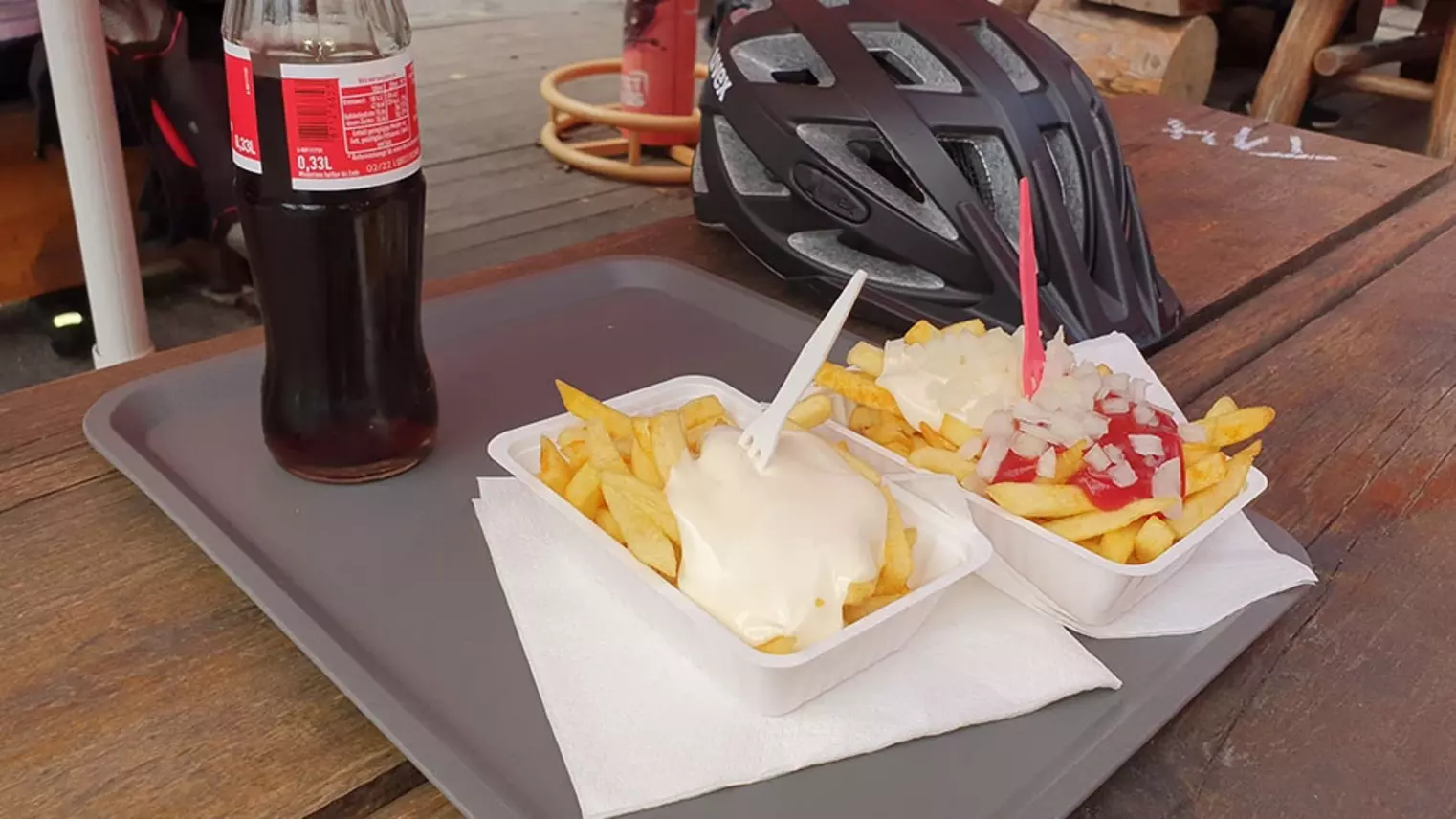 Auf einem Tisch stehen zwei Schalen Pommes mit Mayo und Pommes Spezial sowie eine Flasche Cola. Auch ein Fahrradhelm liegt auf dem Holztisch.