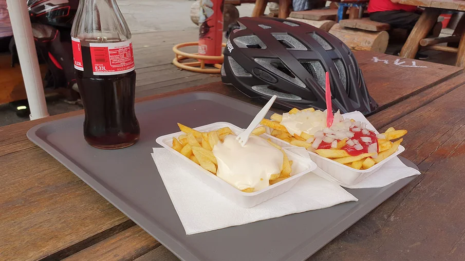 Auf einem Tisch stehen zwei Schalen Pommes mit Mayo und Pommes Spezial sowie eine Flasche Cola. Auch ein Fahrradhelm liegt auf dem Holztisch.