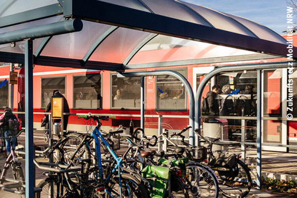 An einem Bahnsteig steht ein roter Zug, Fahrgäste steigen ein, im Vordergrund stehen Fahrräder unter einem Dach