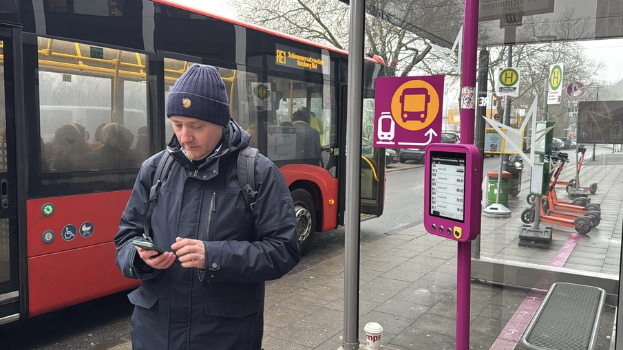 Ein Mensch schaut auf ein Smartphone, im Hintergrund ist ein SEV-Bus und die dazugehörige Haltestelle zu sehen