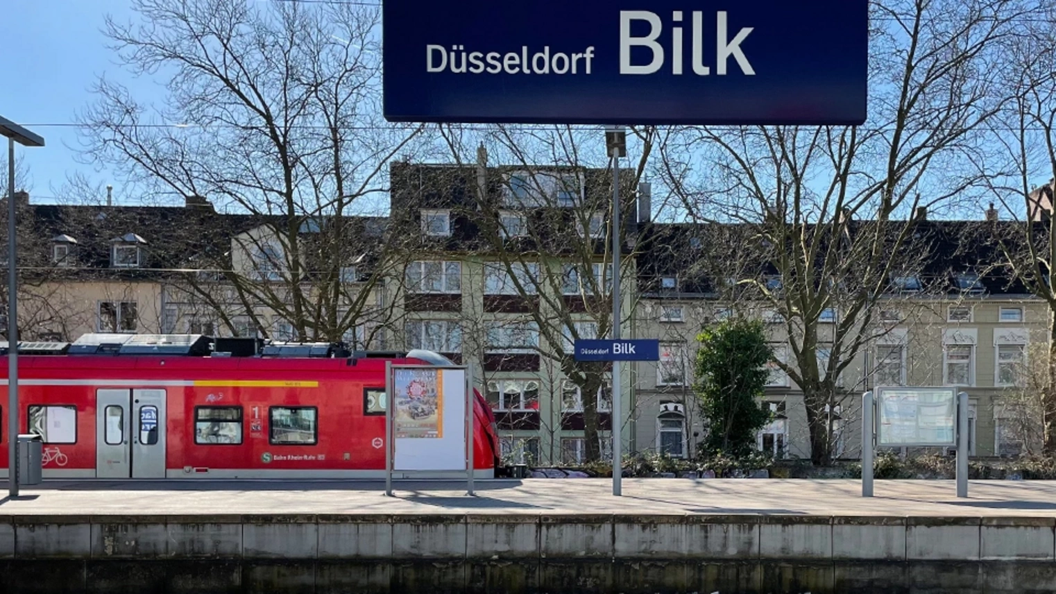 Blick vom Regionalbahnsteig auf den S-Bahn-Bahnsteig mit Zug im Hintergrund, oben im Bild das Stationsschild "Düsseldorf Bilk"
