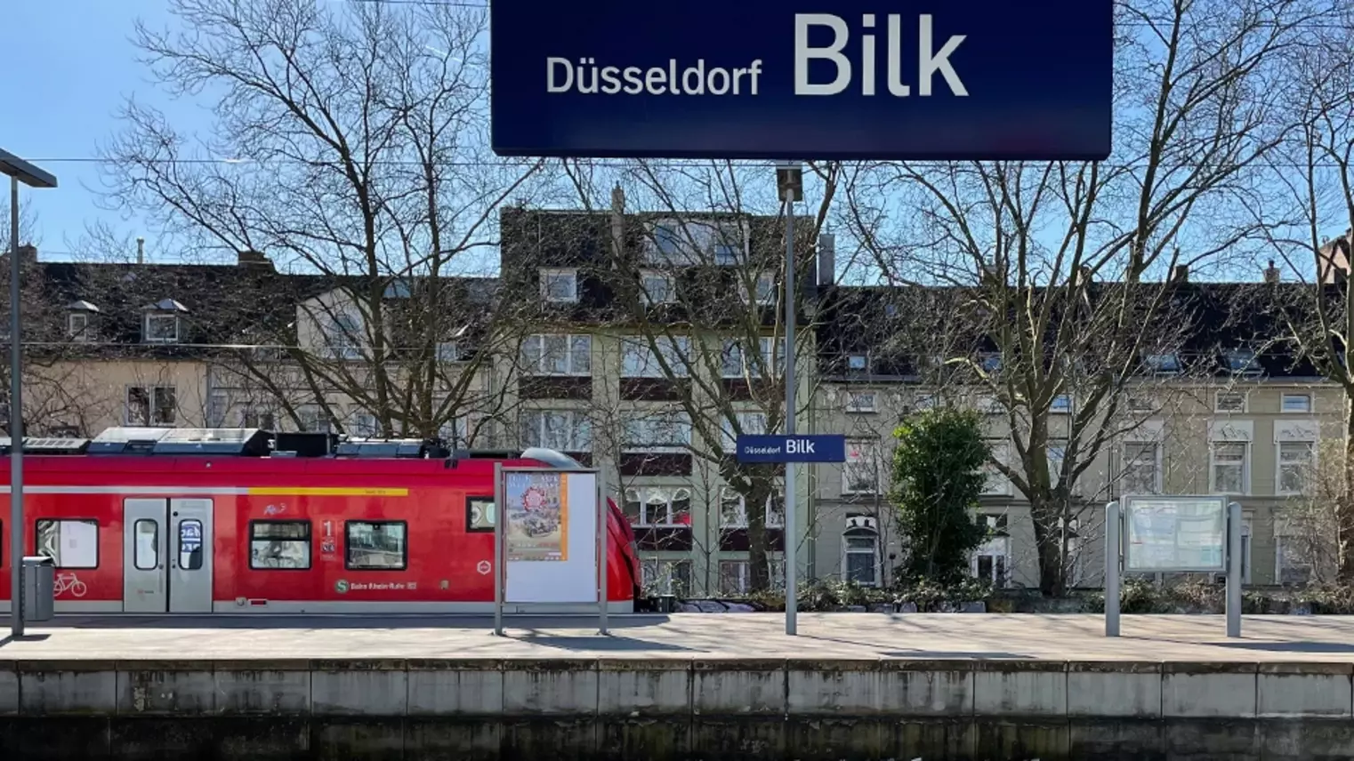 Blick vom Regionalbahnsteig auf den S-Bahn-Bahnsteig mit Zug im Hintergrund, oben im Bild das Stationsschild "Düsseldorf Bilk"