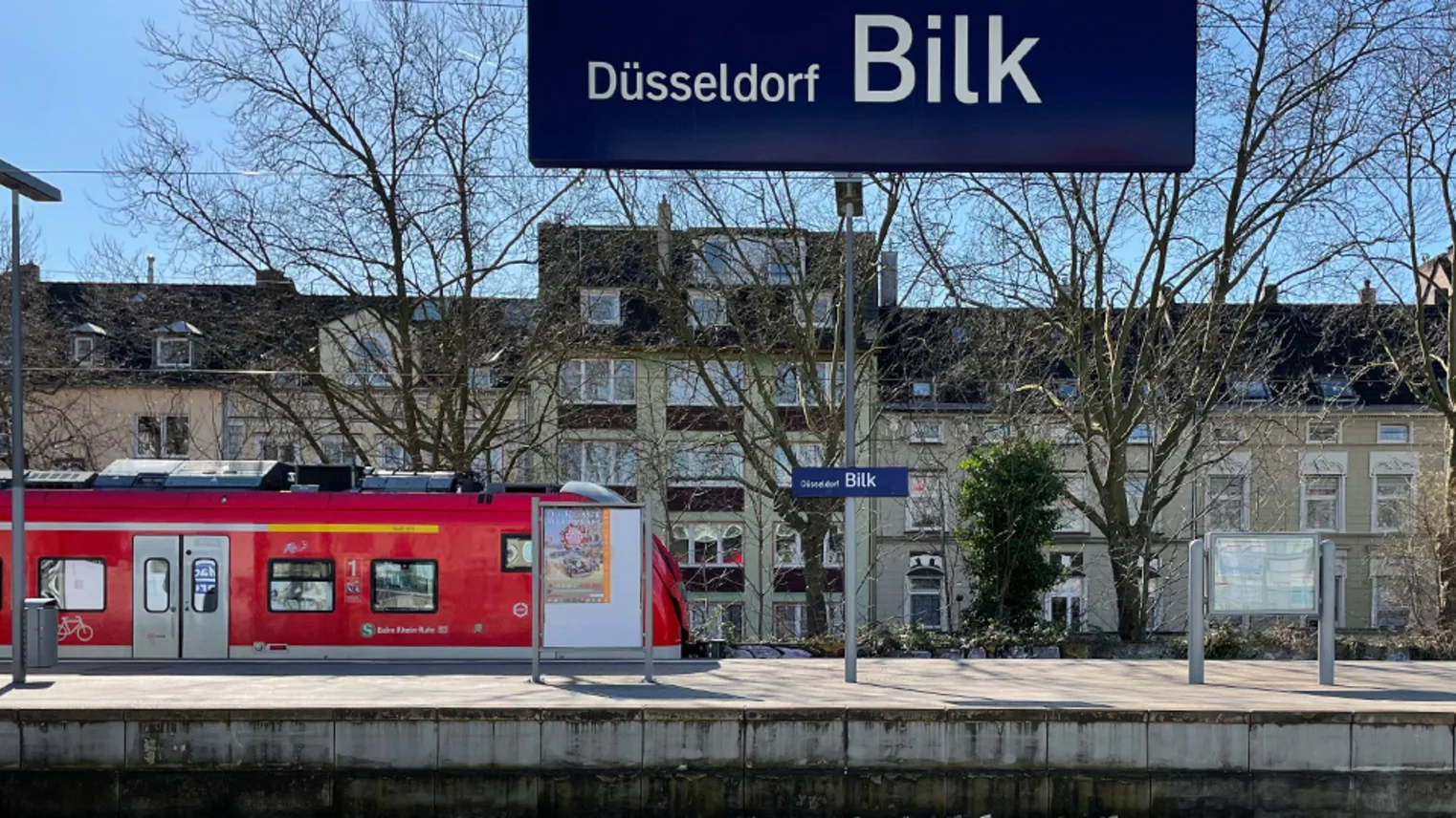 Blick vom Regionalbahnsteig auf den S-Bahn-Bahnsteig mit Zug im Hintergrund, oben im Bild das Stationsschild "Düsseldorf Bilk"