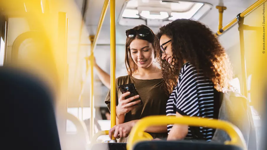 Zwei junge Frauen stehen in einem Bus und blicken auf ein Smartphone