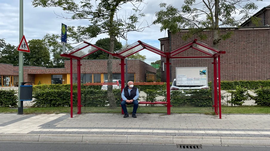 Frontalansicht einer modernisierten Bushaltestelle mit älterem Fahrgast mit Maske