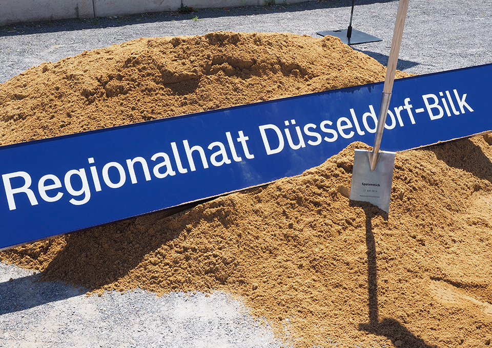 Das Bahnhofsschild von Düsseldorf-Bilk liegt im Sand, daneben ein Spaten