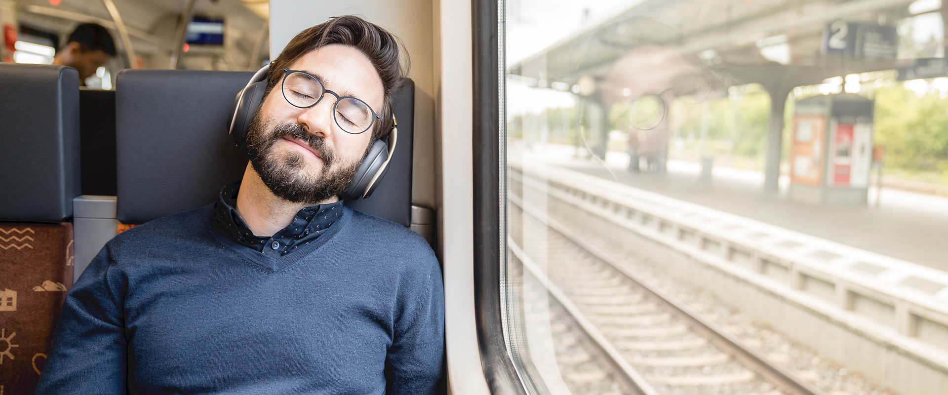Ein junger Mann entspannt mit geschlossene Augen und Kopfhörern auf den Ohren im Zug