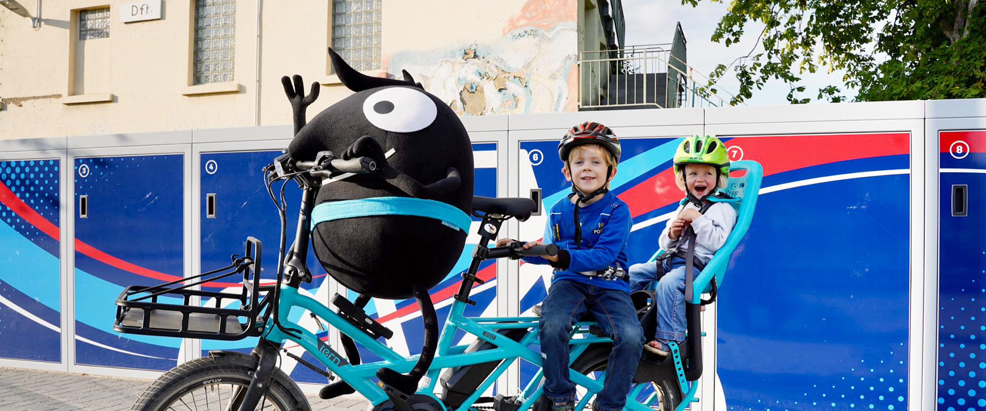 Das Maskottchen der Europäischen Mobilitätswoche und zwei Kinder sitzen gemeinsam auf einem Tandem