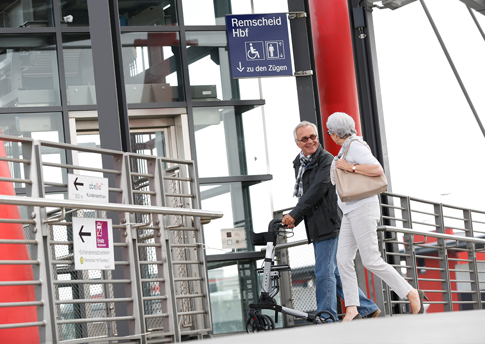 Ein älterer Fahrgast mit Rollator und seine weibliche Begleitung gehen am Bahnhof auf einen Aufzug zu