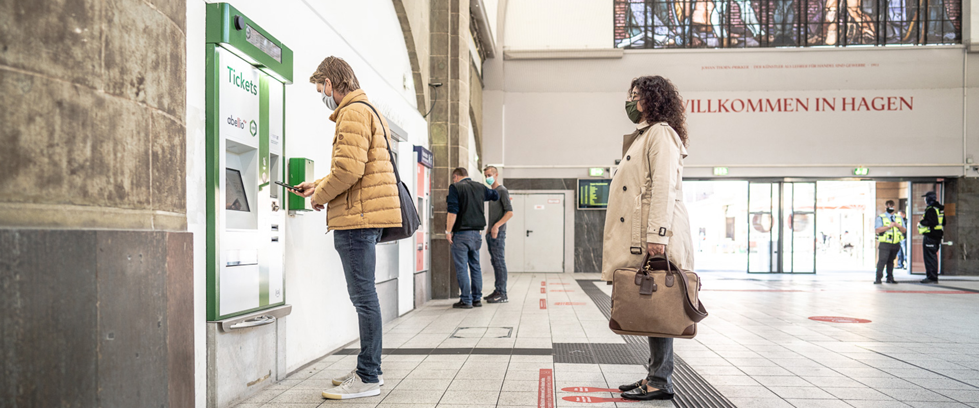 Ein Mann steht in einer Bahnhofshalle an einem Ticketautomaten. Hinter ihm wartet eine Frau, die sich auch ein Ticket kaufen möchte.