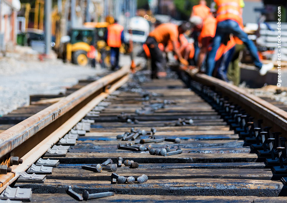 Arbeiten an Schienen, im Hintergrund sind unscharf Bauarbeiter*innen und Baumaschinen zu sehen