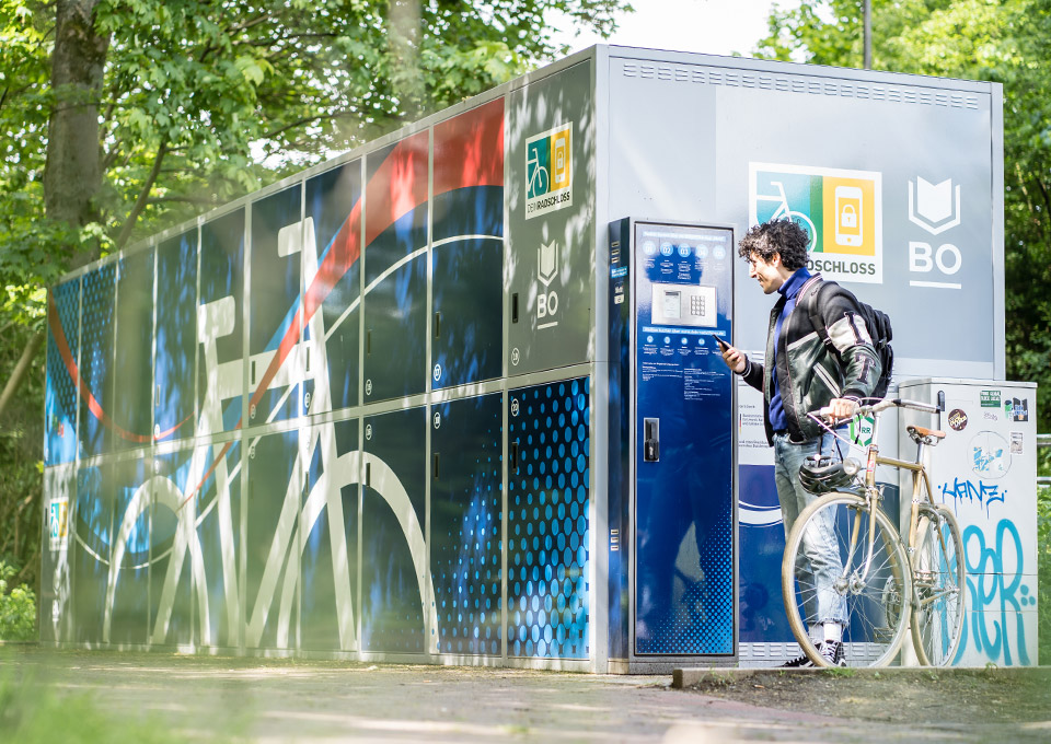 Ein junger Mann steht mit seinem Fahrrad an den Radboxen von Dein Radschloss