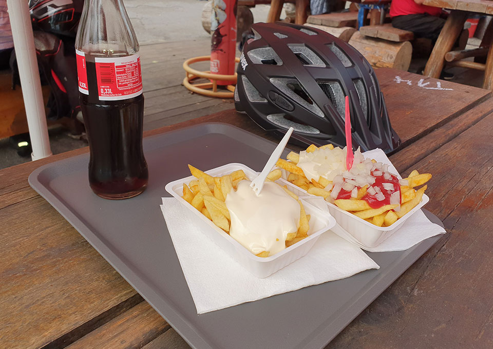 Auf einem Tisch stehen zwei Schalen Pommes mit Mayo und Pommes Spezial sowie eine Flasche Cola. Auch ein Fahrradhelm liegt auf dem Holztisch.
