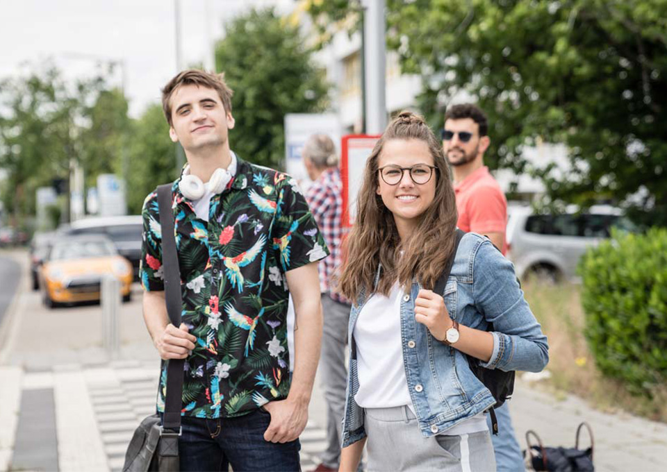 Ein Mädchen und ein Junge stehen gemeinsam an der Bushaltestelle und schauen zu dem heranfahrenden Bus.