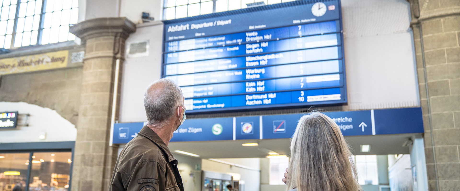 Zwei Personen stehen in einer Bahnhofshalle und blicken auf eine Abfahrtstafel