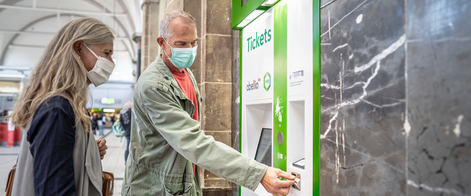 Ein Mann und eine Frau, die eine Gesichtsmaske tragen, stehen am Ticketautomat und bezahlen ein Ticket