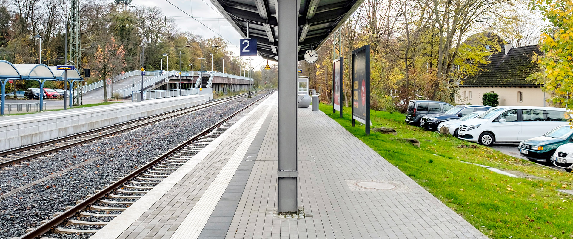 Blick auf einen überdachten Bahnsteig, links Gleise, rechts Autos auf einem P+R-Parkplatz