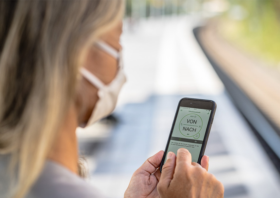 Frau mit Maske blickt auf die Verbindungssuche in der VRR App auf ihrem Smartphone