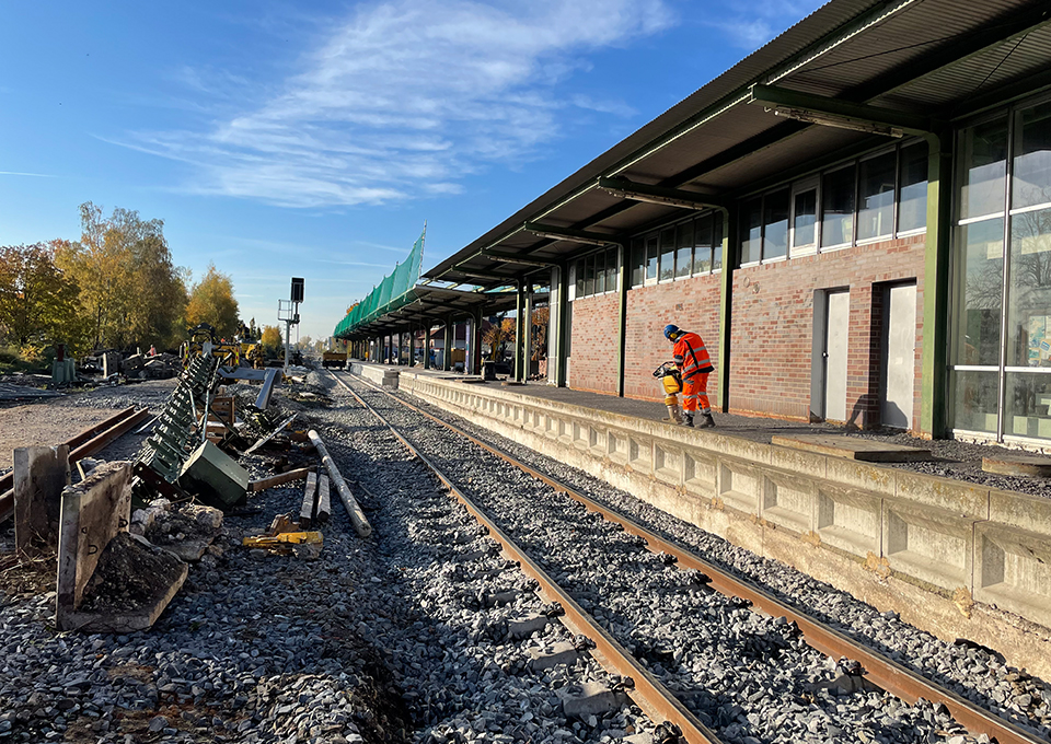 Ansicht eines Bahnsteigs mit Gleisen davor, an dem ein Bauarbeiter mit einem Vibrationsstampfer der Boden verdichtet
