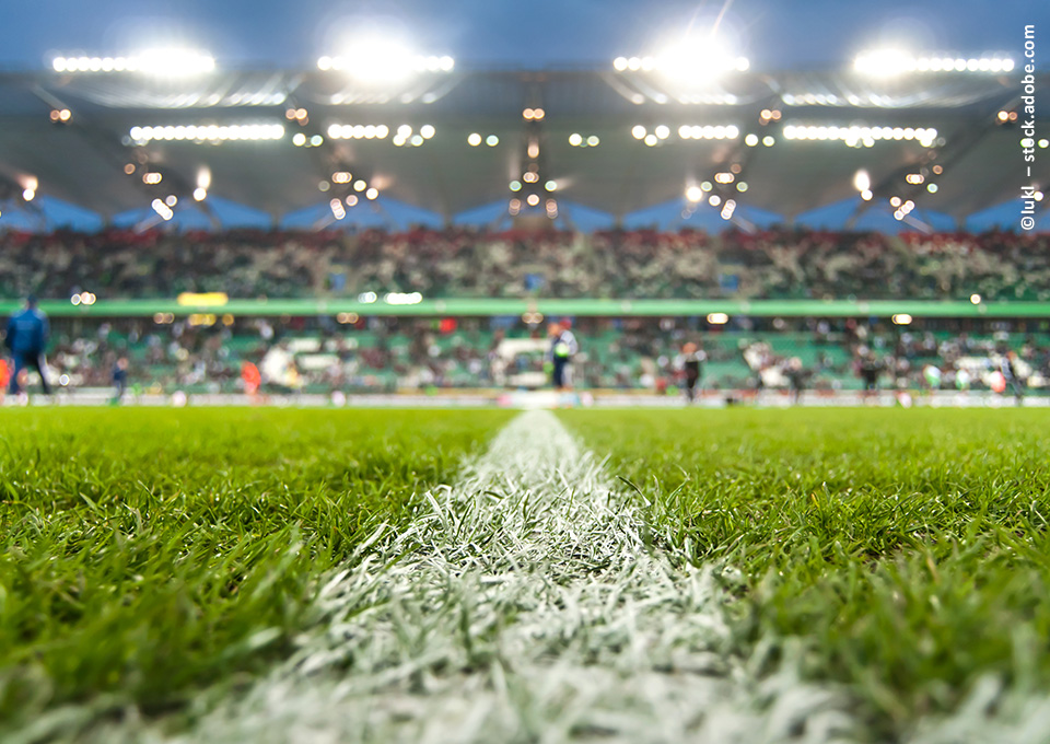 Der Fußballrasen und die Mittellinie stehen im Fokus des Bildes. Im Hintergrund ist eine Zuschauertribüne zu erkennen.