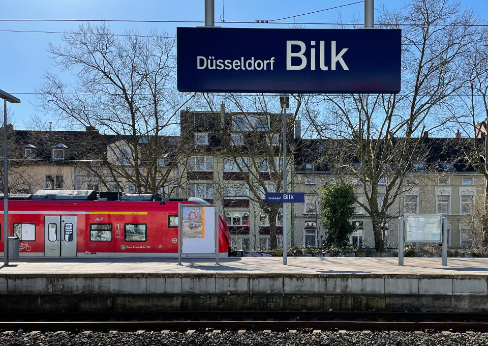 Blick vom Regionalbahnsteig auf den S-Bahn-Bahnsteig mit Zug im Hintergrund, oben im Bild das Stationsschild "Düsseldorf Bilk"