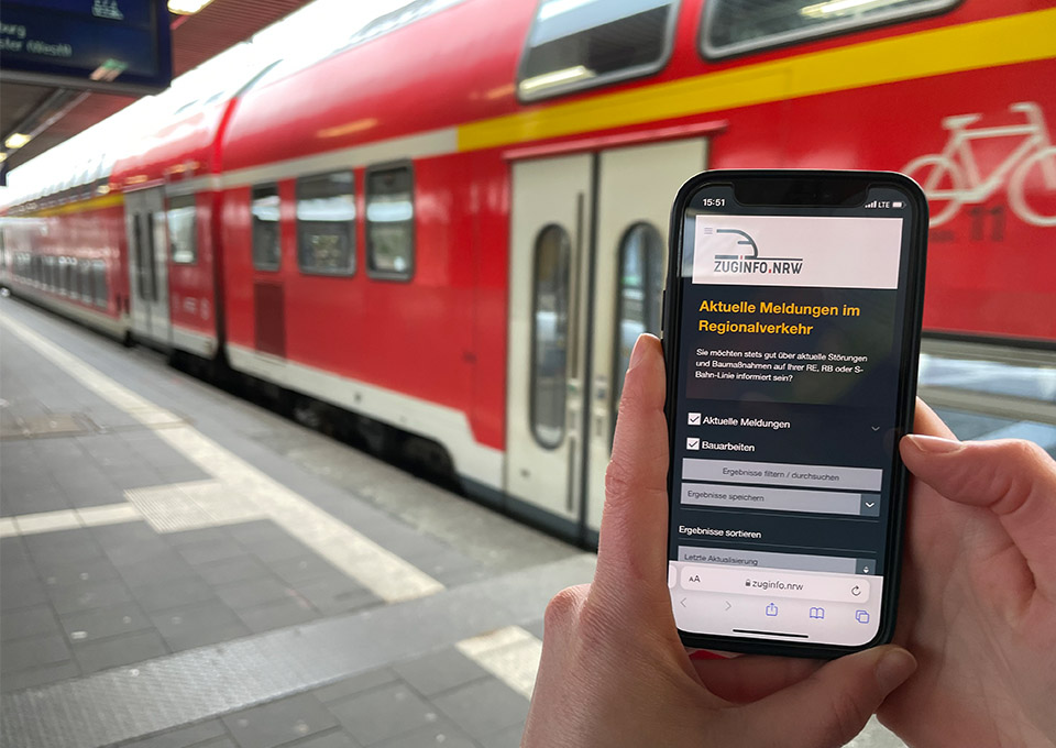 Eine Person am Bahnsteig, im Hintergrund ein roter Zug, hält ein Handy in der Hand. Im Display 