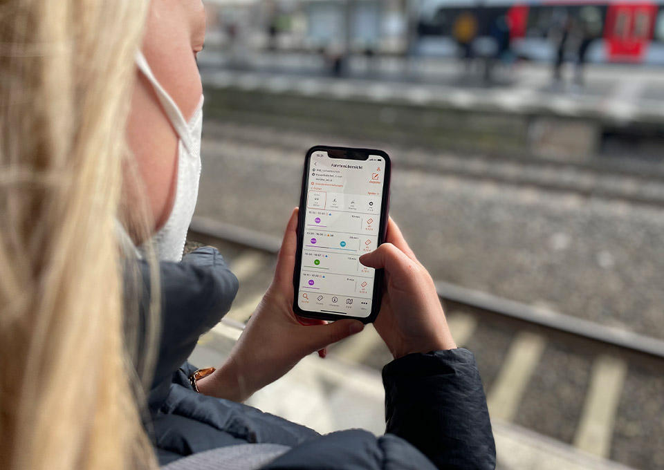 Blick über die Schulter einer Person auf ein Smartphone. Im Display zu sehen ist die Fahrplanauskunft in der VRR App.