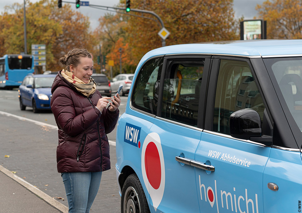 Eine junge Frau steht vor einem "WSW Cabs"-Fahrzeug und tippt auf ihr Smartphone